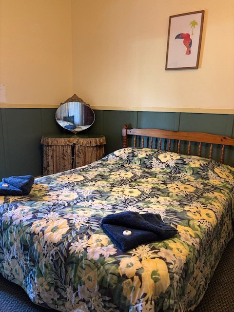 A bedroom with a wooden bed frame, floral bedspread, a small rustic wooden dresser with a round mirror, a wall decoration of a parrot, leaf, and toucan, and two folded towels on the bed.