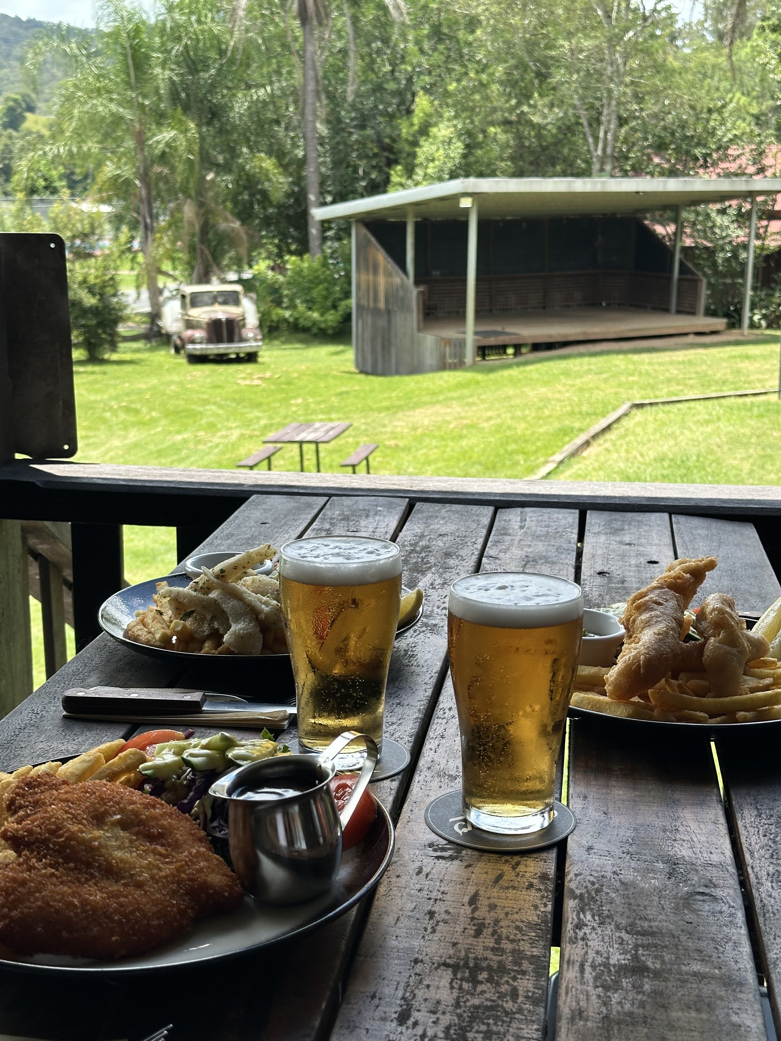 Outdoor table with two glasses of beer, fried fish, French fries, salad, and coleslaw on a rustic wooden table, with a grassy yard, picnic table, and a covered stage in the background.