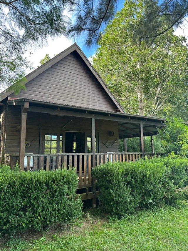 A wooden house with a front porch, surrounded by green bushes and trees, under a partly cloudy sky.
