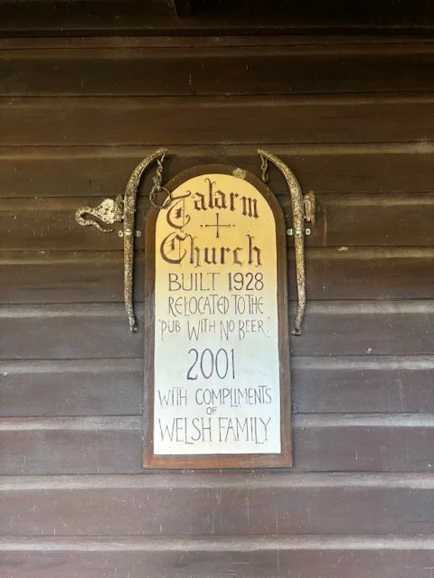 Wooden sign with decorative horseshoes, indicating the church was built in 1928, relocated to the pub with no beer in 2001, and given with compliments of the Welsh family.