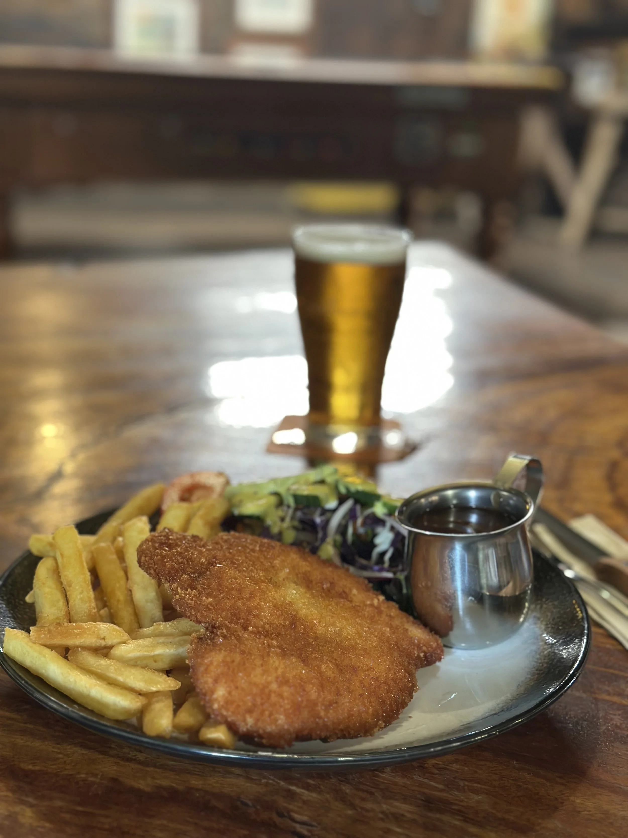 Plate with breaded fried fish, French fries, salad with dressing, and a small silver pitcher of tartar sauce, with a glass of beer in the background.