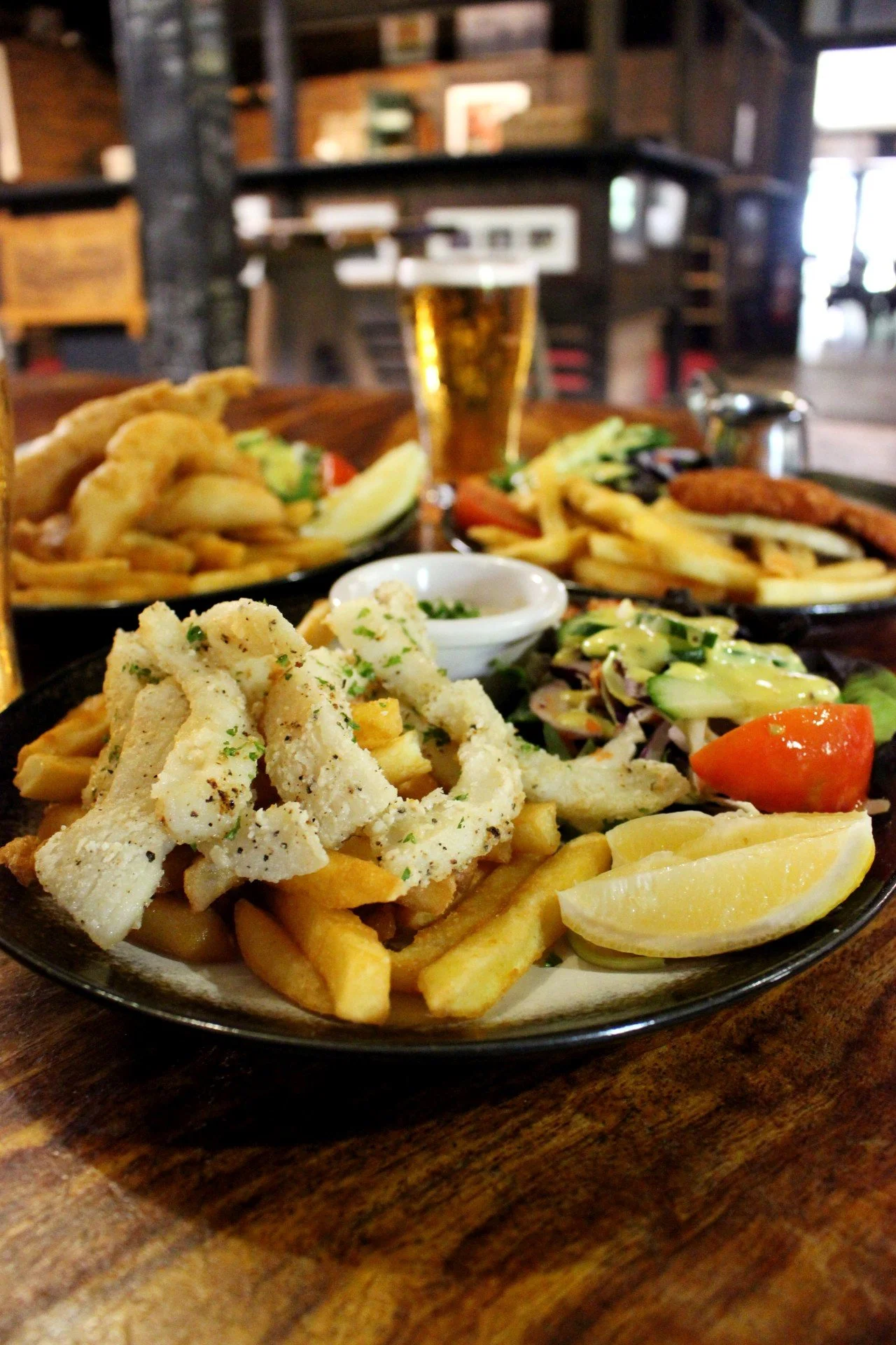 Plate of fried calamari with fries, lemon wedges, and a side salad, with a pint of beer in the background on a wooden table inside a restaurant.