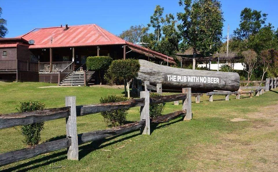 A large wooden log with the text 'THE PUB WITH NO BEER' engraved on it, situated on a grassy area with a wooden fence and a house with a red roof in the background.