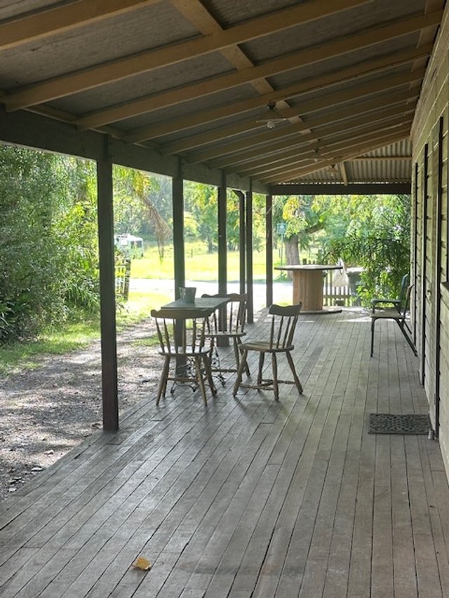 Covered outdoor porch with wooden flooring, a small round table with four chairs, and greenery in the background.