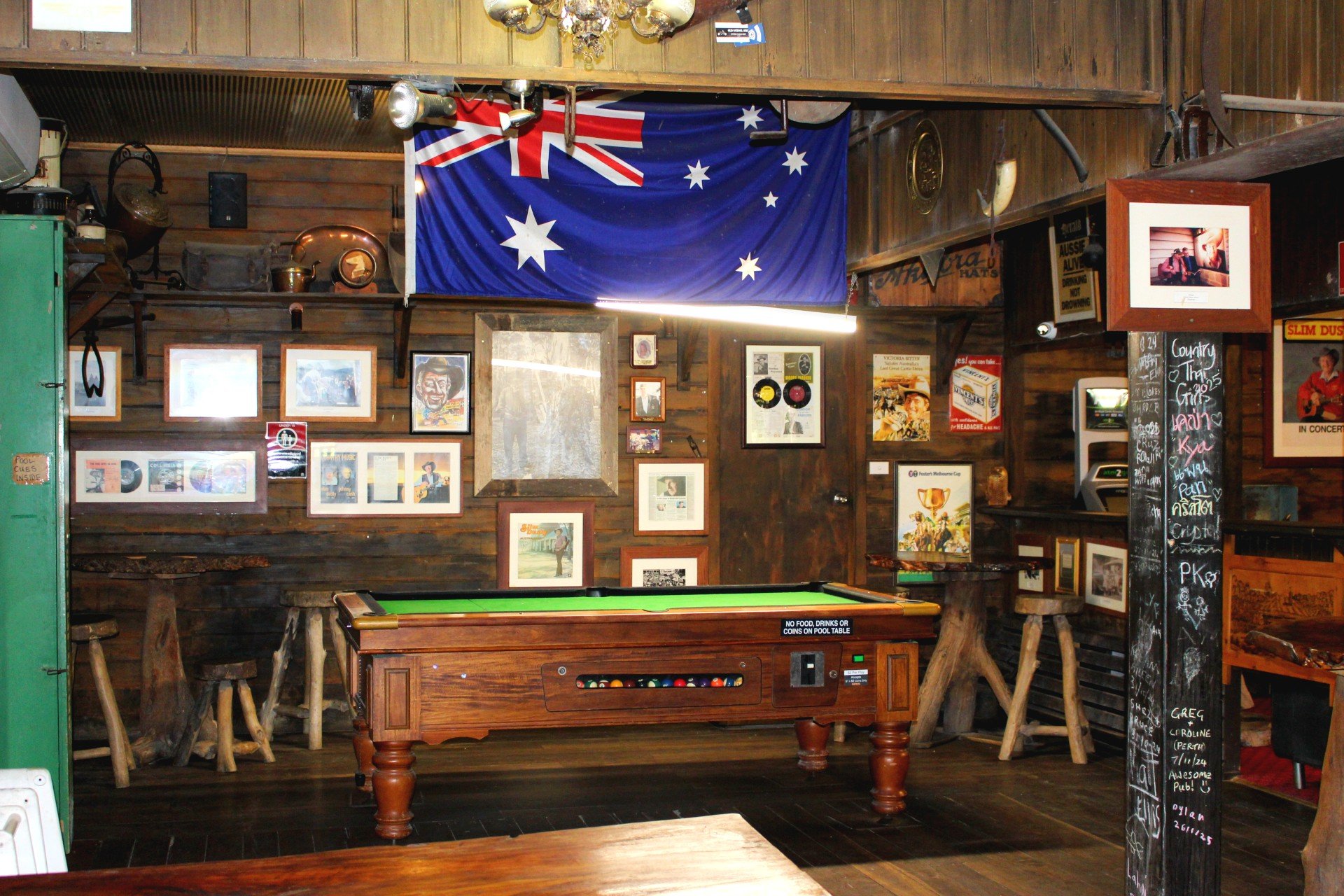 Interior of a rustic pub or bar with wooden walls decorated with framed pictures, a large Australian flag hanging from the ceiling, a pool table, a chalkboard with handwritten notes, and stools around the room.