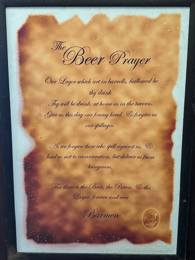 A framed paper titled "The Beer Prayer" written in script. It is an old parchment style with burned edges and a small logo at the bottom right corner. The prayer humorously seeks blessings for beer.
