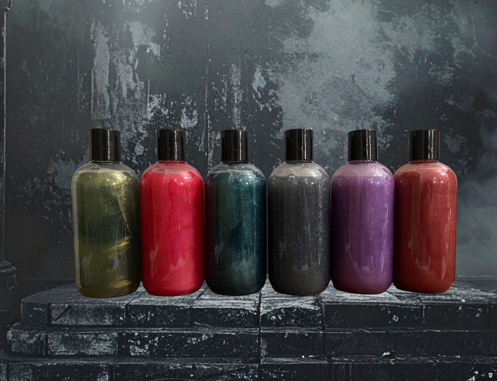 Six bottles of paint with black caps, in colors olive green, red, teal, black, purple, and red-orange, placed on a stone surface with a dark textured background.