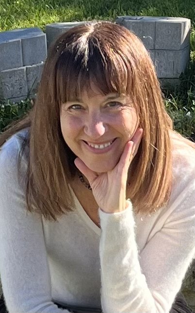 A woman with shoulder-length reddish hair smiling and resting her chin on her hand outdoors with grass and concrete blocks in the background.