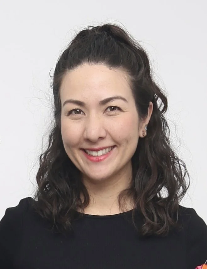 A young woman with dark curly hair tied back, smiling, wearing a black top and small earrings, in front of a plain white background.