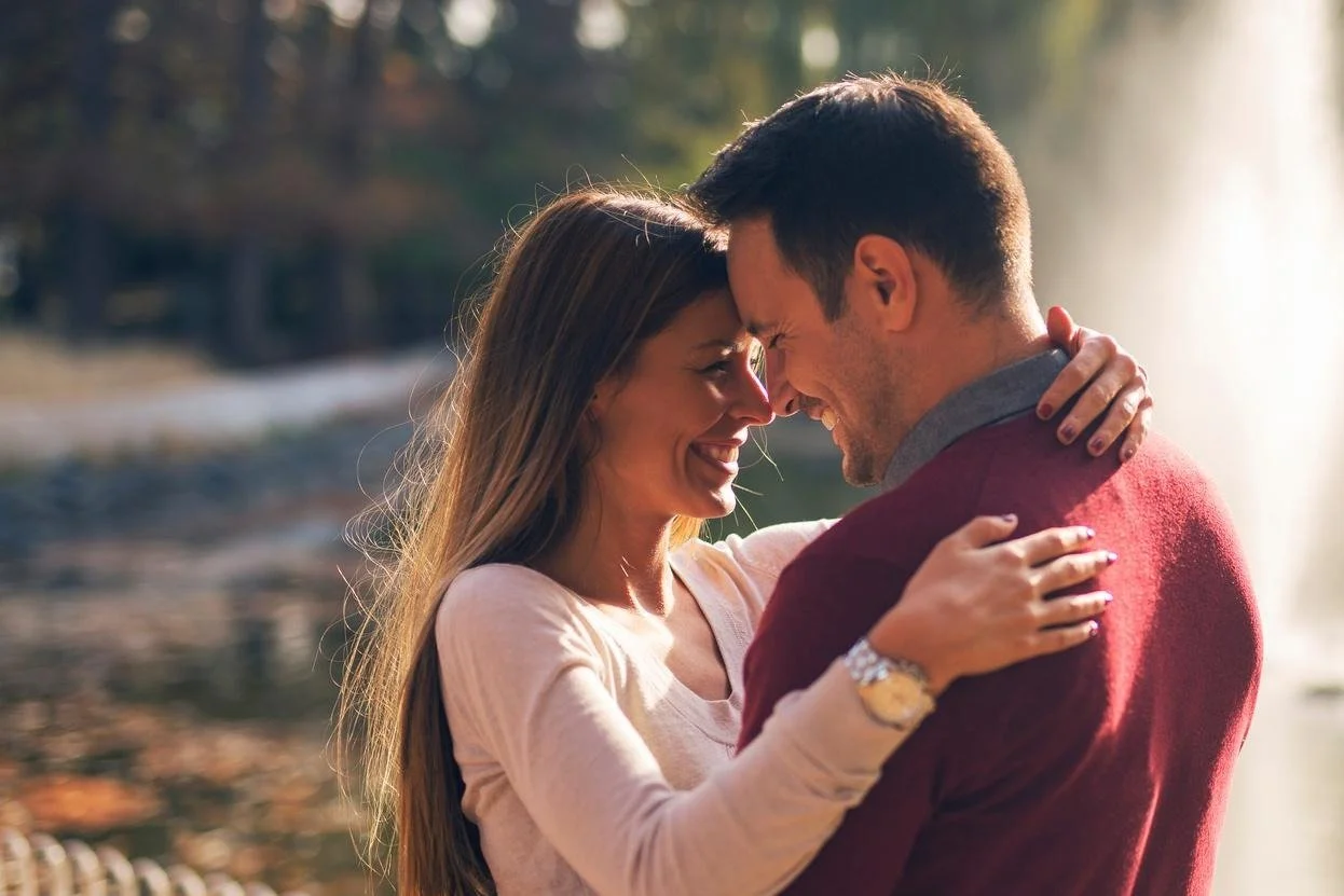 A couple smiling and touching foreheads outdoors near a body of water with trees in the background