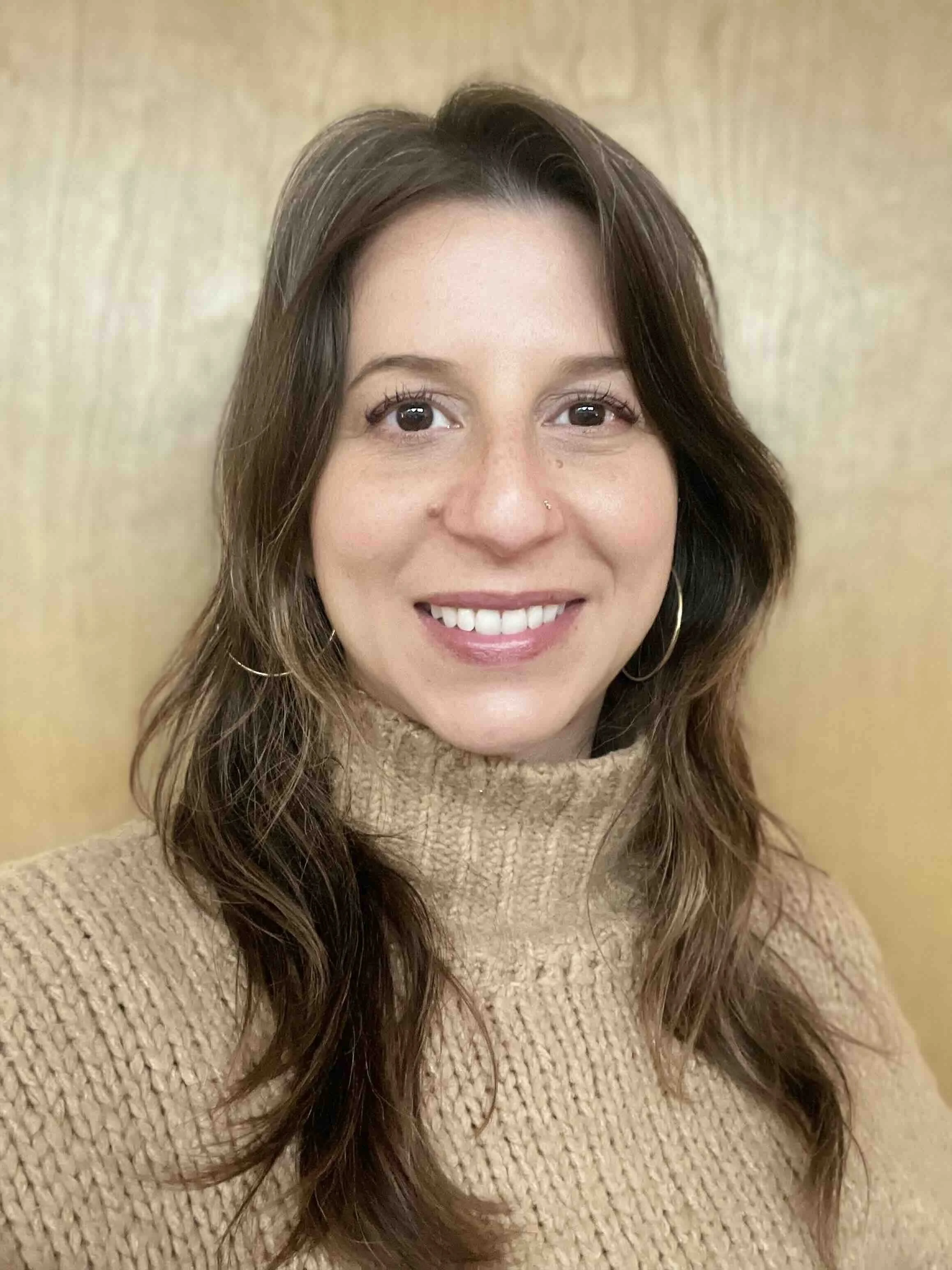 A woman with long brown wavy hair, wearing a beige turtleneck sweater and hoop earrings, smiling at the camera against a beige background.