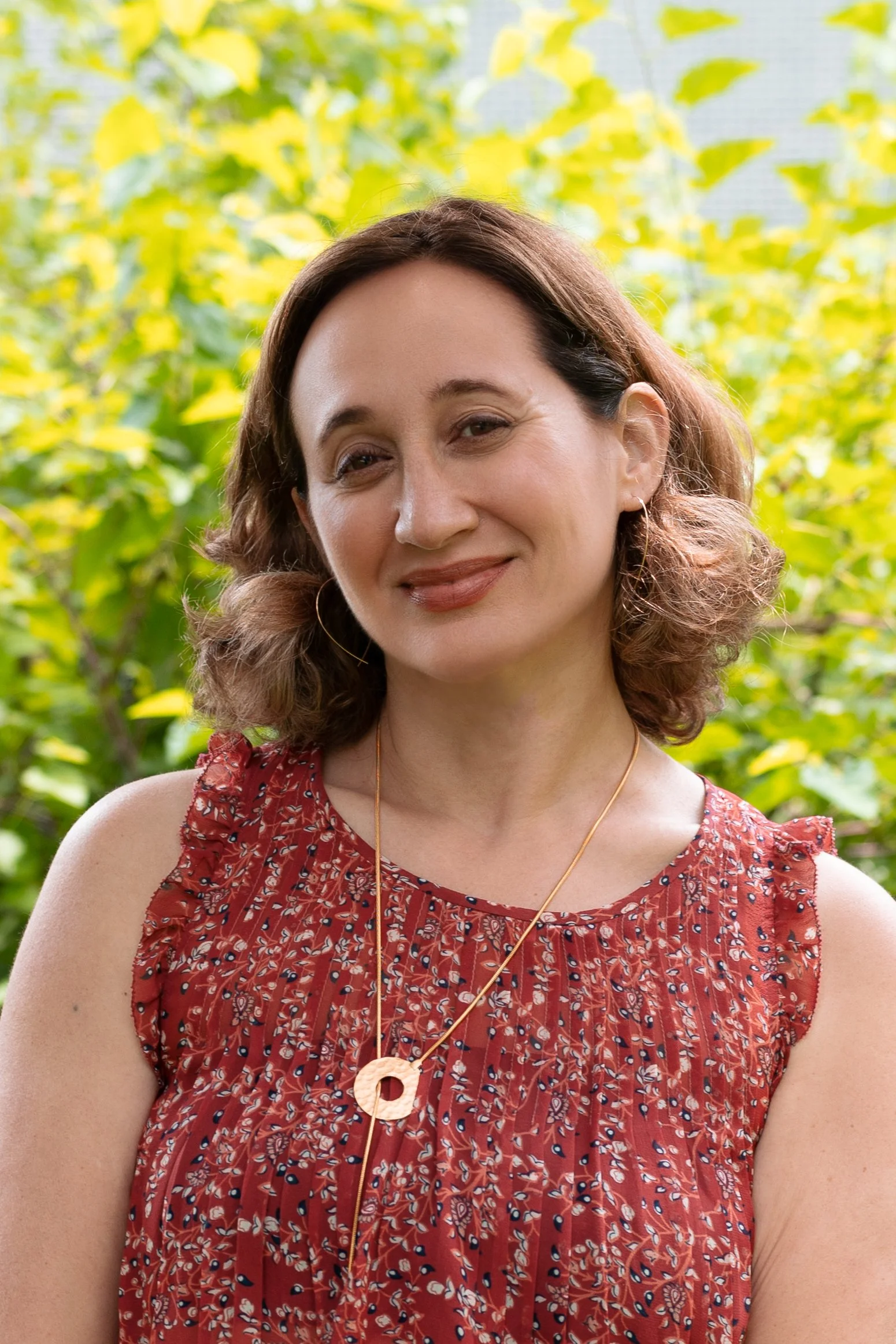 A woman with shoulder-length wavy brown hair, wearing a red sleeveless dress with a floral pattern, standing outdoors with green foliage in the background. She is smiling gently and wearing a gold necklace with a round pendant and hoop earrings.