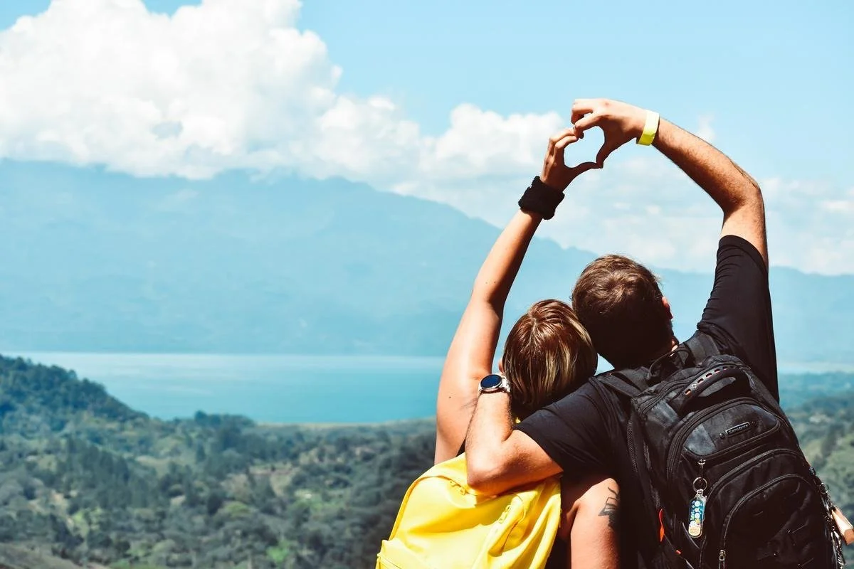 A couple with backpacks forming a heart shape with their raised arms against a scenic mountain and lake backdrop.