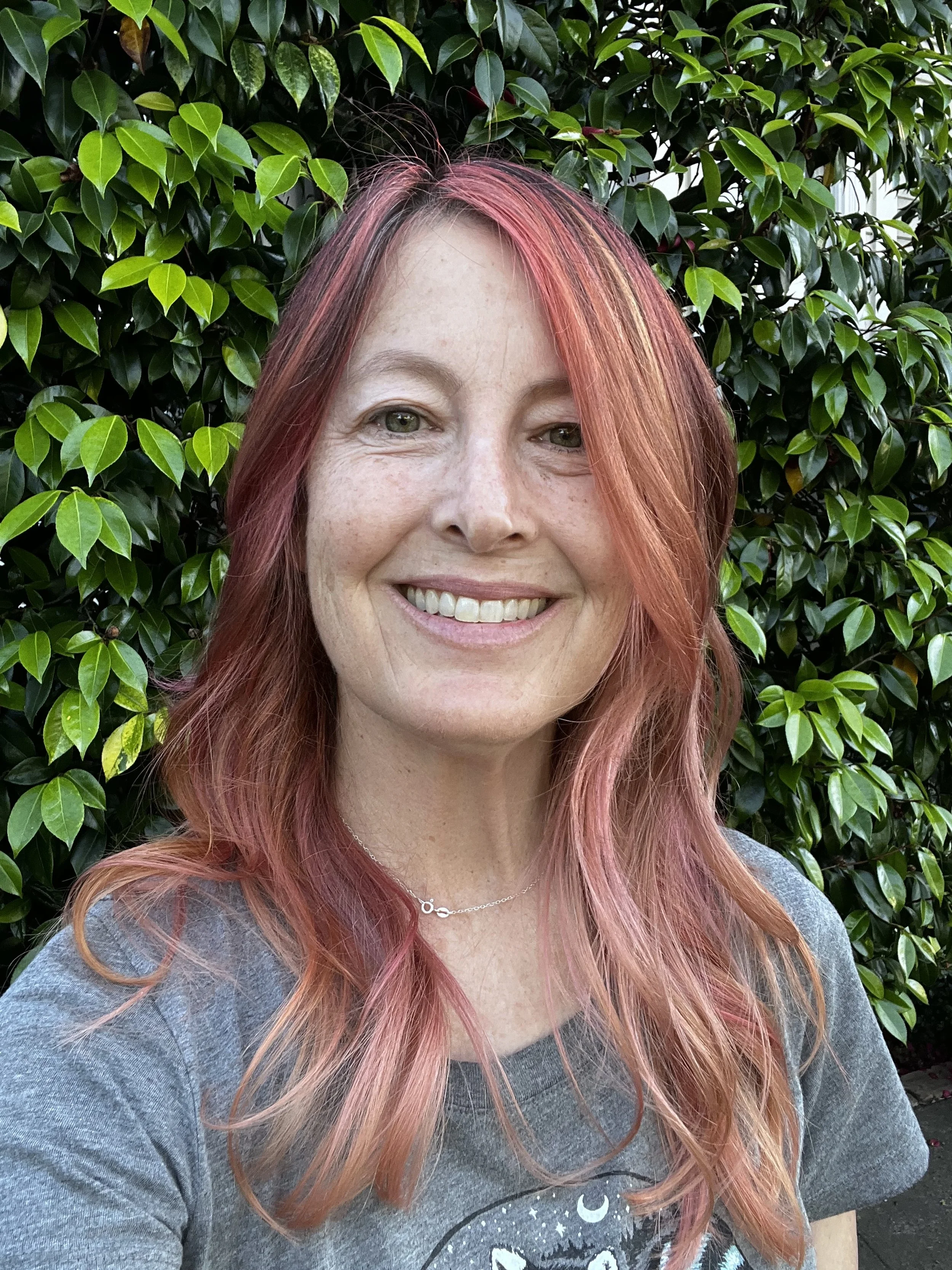 A woman with reddish-pink hair smiling outdoors in front of green leafy bushes.