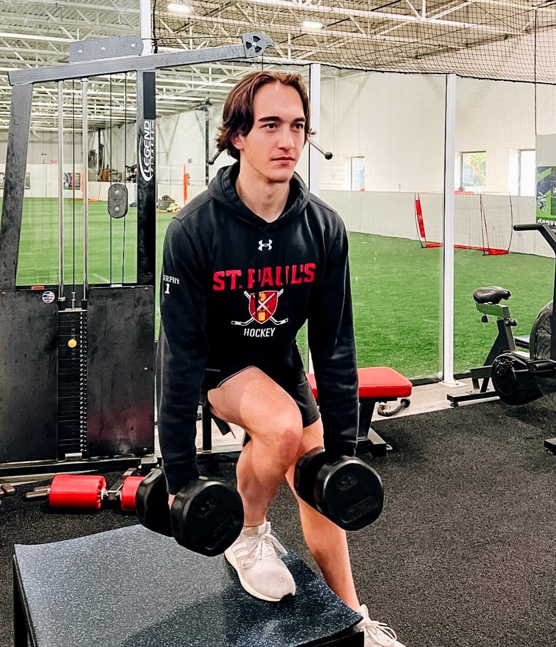 A person wearing a "St. Paul's Hockey" hoodie performs a lunge exercise with dumbbells in a gym environment. The person is on one knee on a platform. Exercise equipment and an indoor sports field are visible in the background.