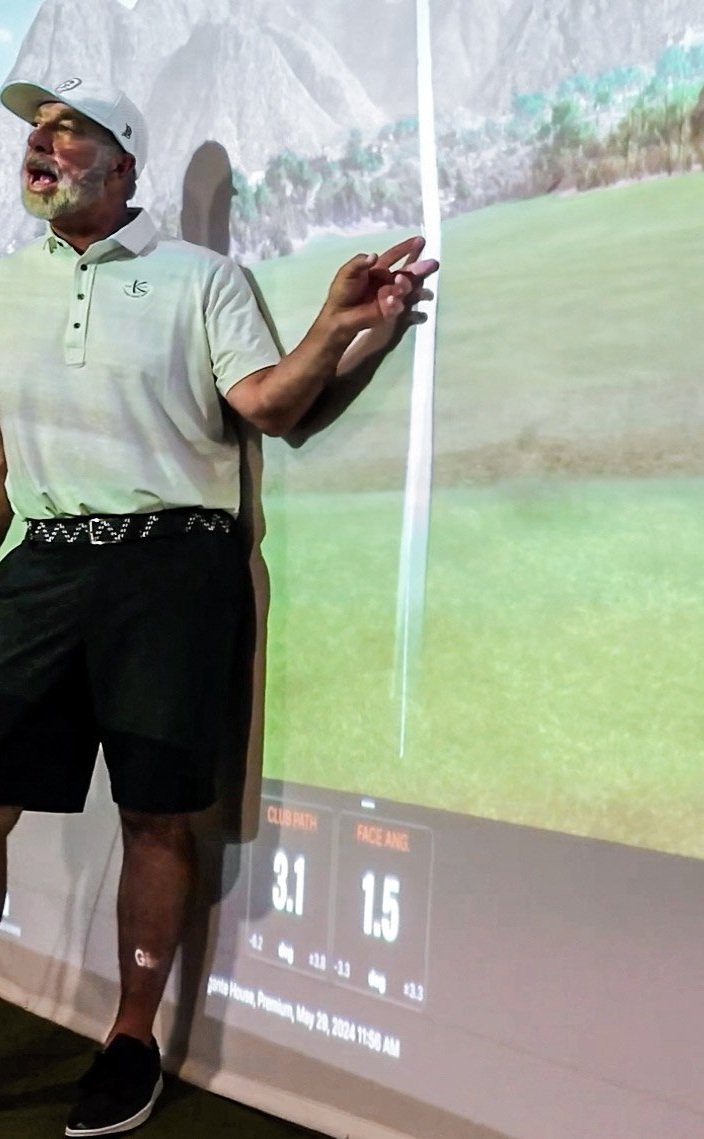 A man wearing a white polo shirt, black shorts, and a white cap standing in front of a golf simulator screen displaying golf swing data. The background features a landscape with mountains and a golf course.