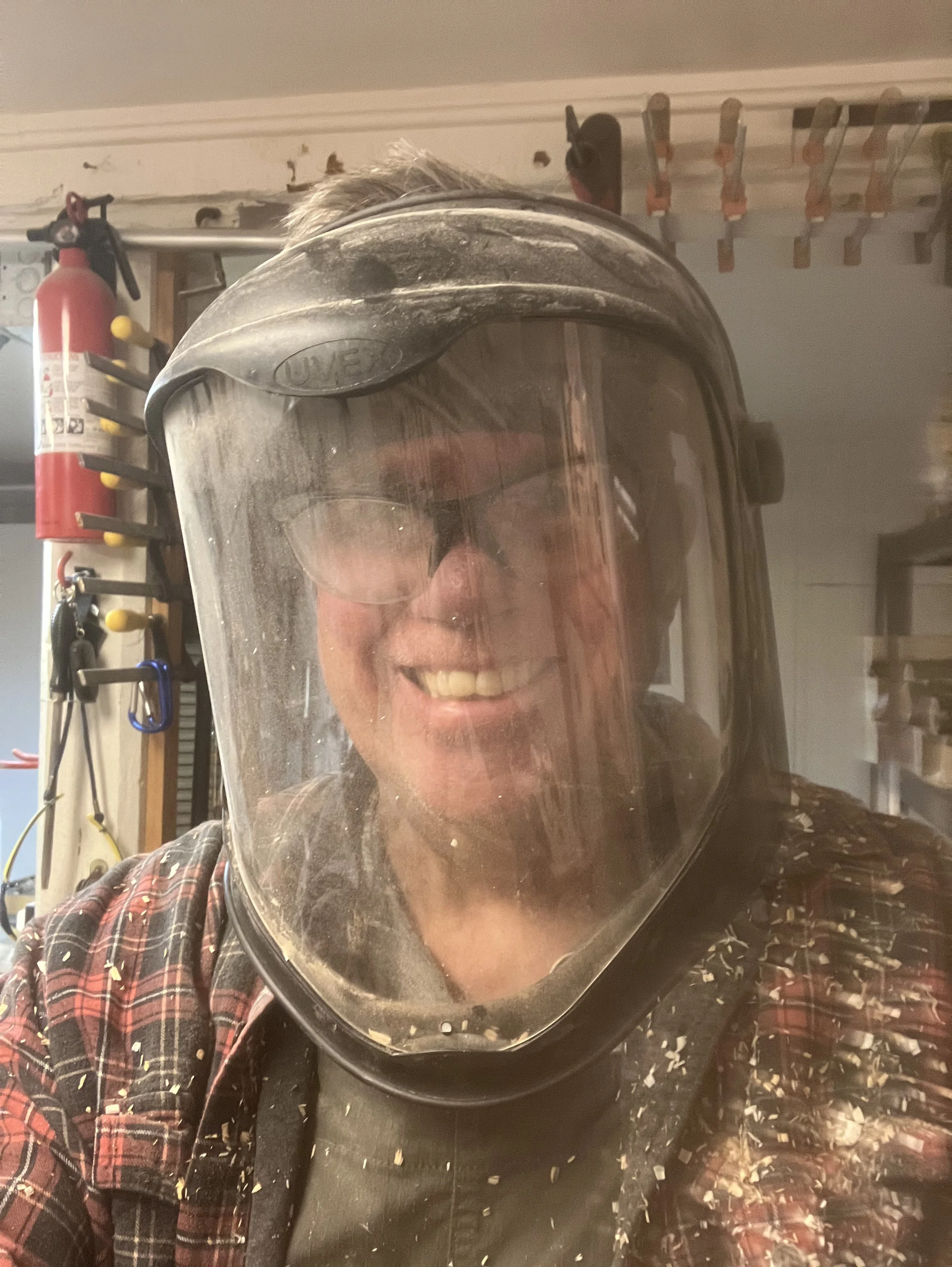 Person wearing a protective face shield in a workshop, covered in sawdust, with tools and a fire extinguisher in the background.