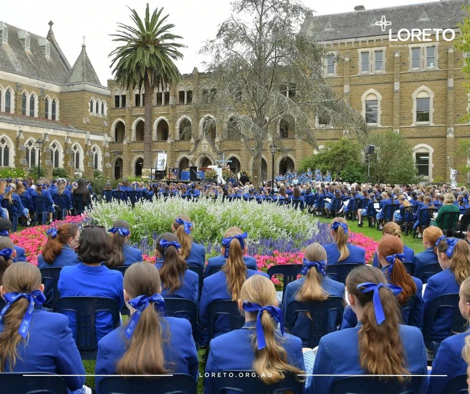 A large group of students in blue uniforms and blue ribbons seated outdoors on green grass, attending an event or ceremony in front of an old stone building with arched windows, surrounded by flowers and trees.
