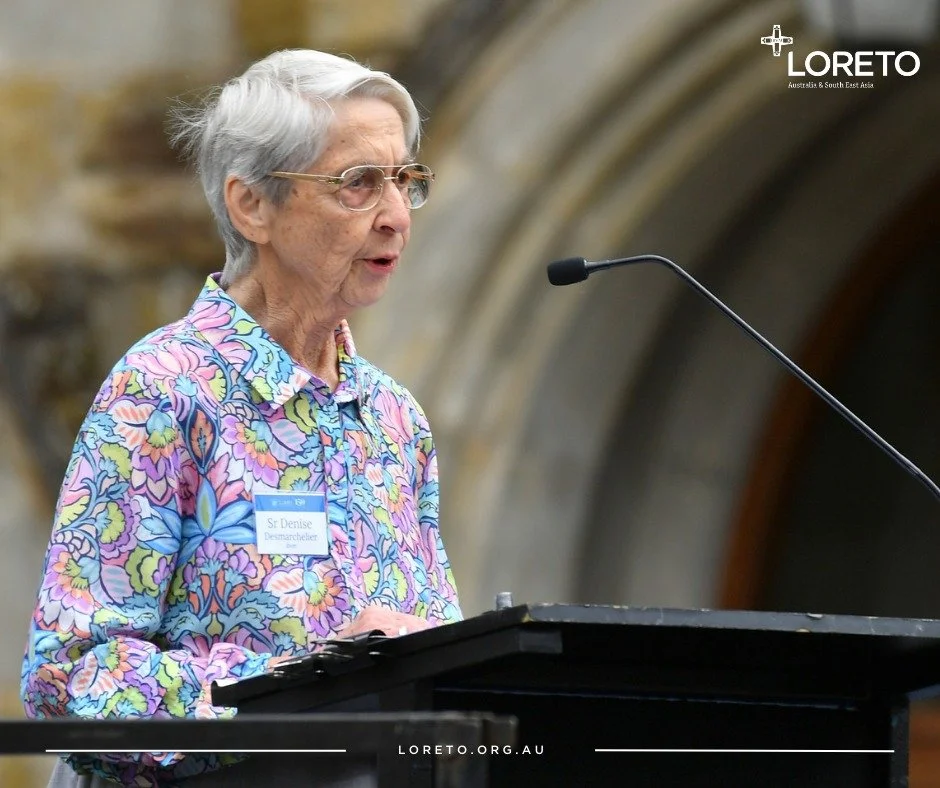 An elderly woman with gray hair, glasses, and a colorful floral shirt speaking at a podium during an event at a church or cathedral. There is a microphone in front of her and a sign on her shirt that reads 'Sr Denise Desmarchelier.' The background sh