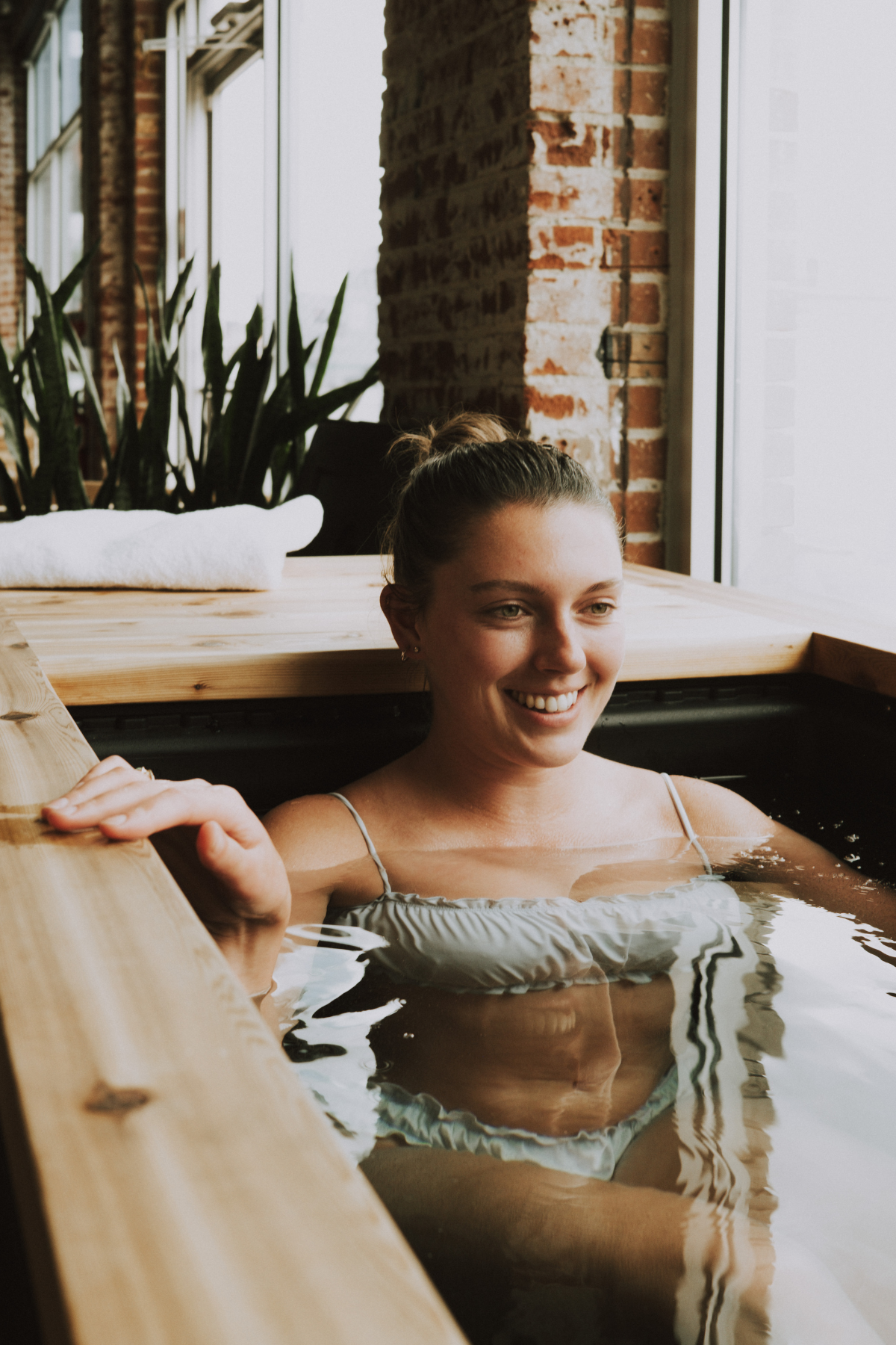 A young woman relaxing in a wooden cold plunge tub, smiling, with water up to her chest, in a room with brick walls and large windows, in a neighborhood wellness club in Denver.