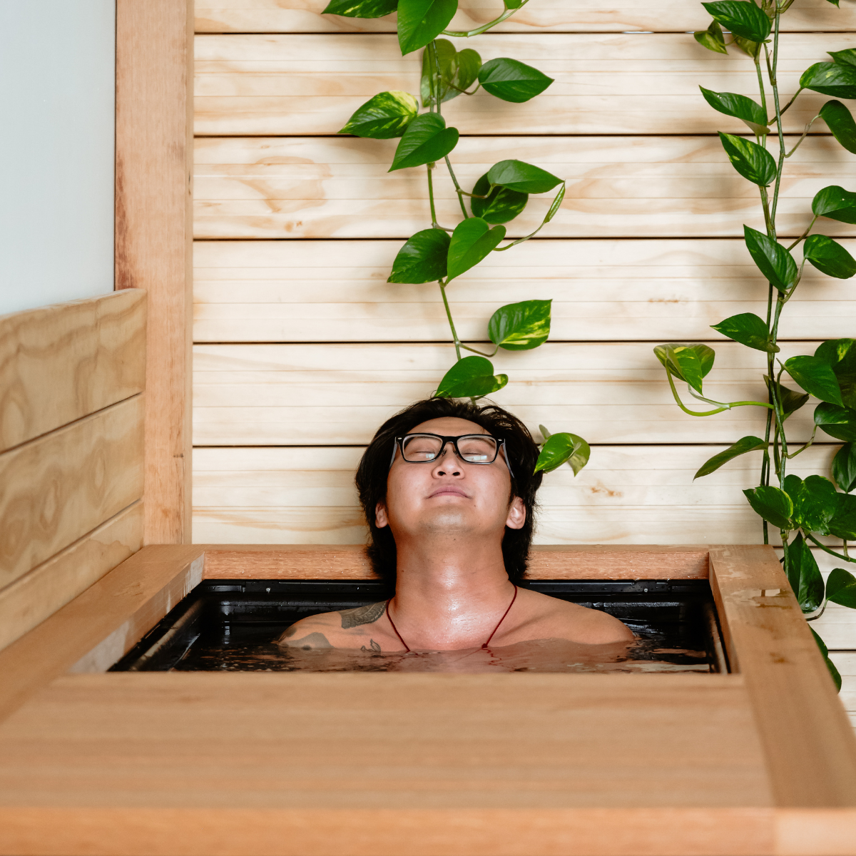 A man with glasses and tattoos relaxing in a cold plunge with closed eyes, surrounded by green leafy plants and wooden wall panels in a neighborhood wellness club in Denver.