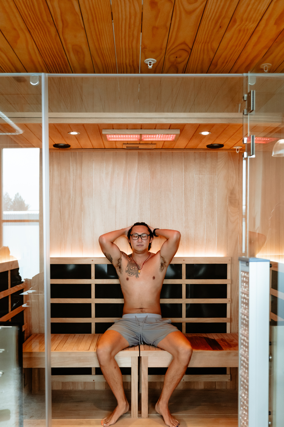 A man with tattoos and glasses sitting shirtless in a wooden infrared and red light sauna, resting with hands on his head and eyes closed, in a neighborhood wellness club in Denver.