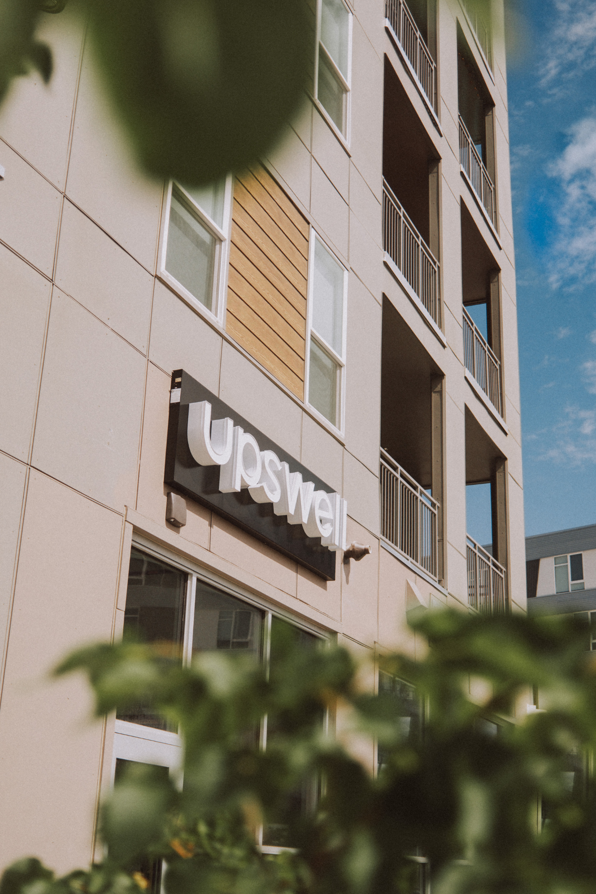 Exterior view of a modern apartment building with a sign that reads 'Upswell'. The building has large windows and balconies, with some greenery partially blocking the view.