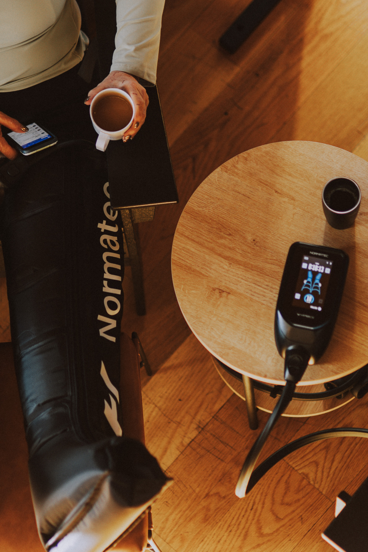 Top-down view of a person sitting at a table with a mug of coffee, a smartphone, and Normatec compression leg sleeves. The person is holding the mug in one hand and is sitting in a neighborhood wellness club in Denver.