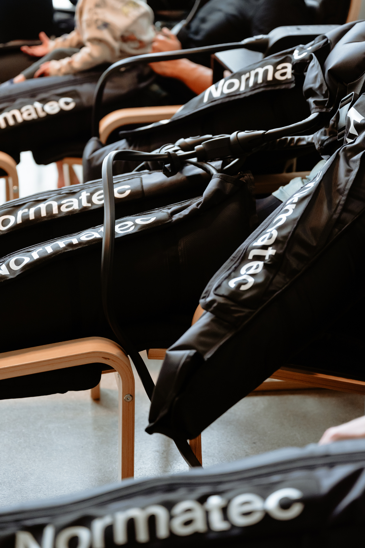 Rows of black leg compression sleeves labeled 'Normatec' on wooden chairs in a waiting area with a person in the background, in a neighborhood wellness club in Denver.