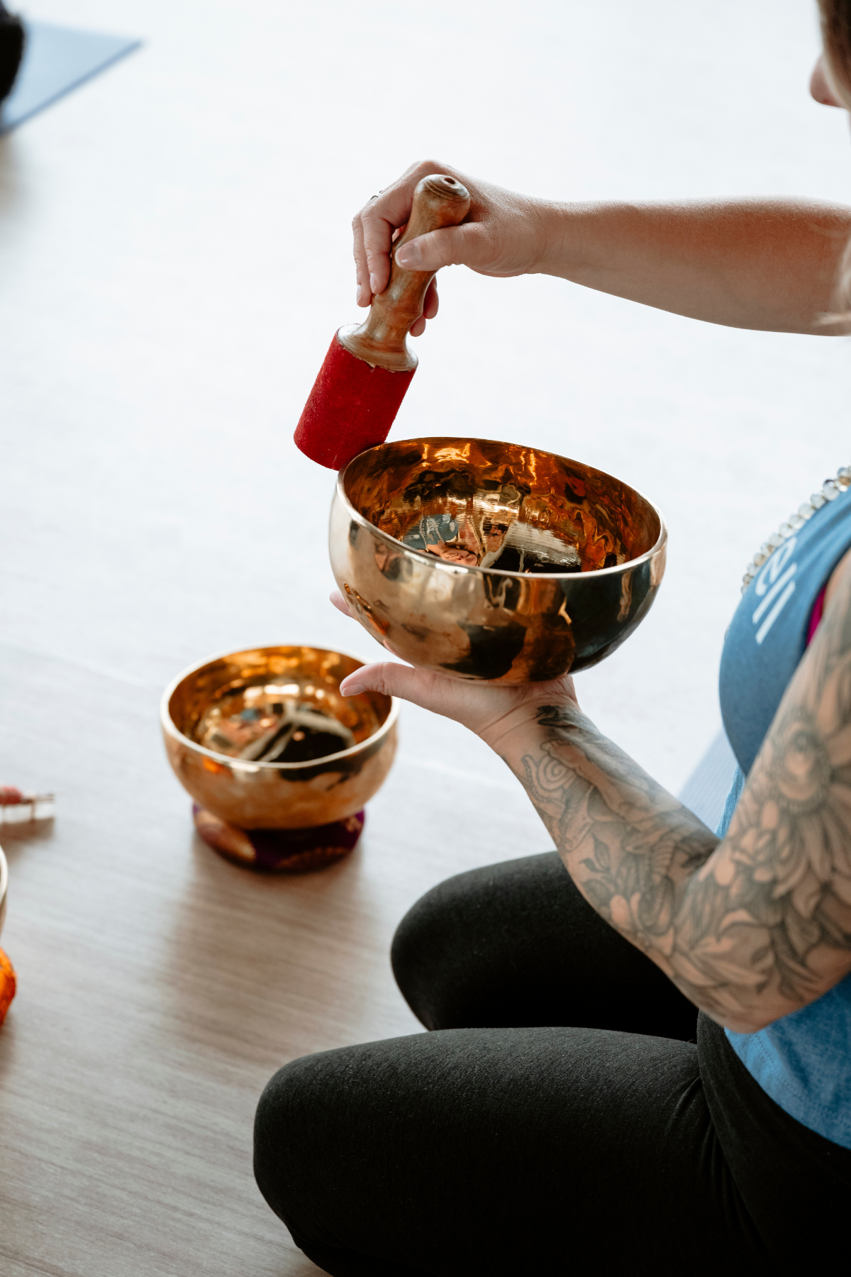 Person with tattooed arm holding a copper singing bowl and a wooden mallet, another singing bowl placed on the table in a neighborhood wellness club in Denver.