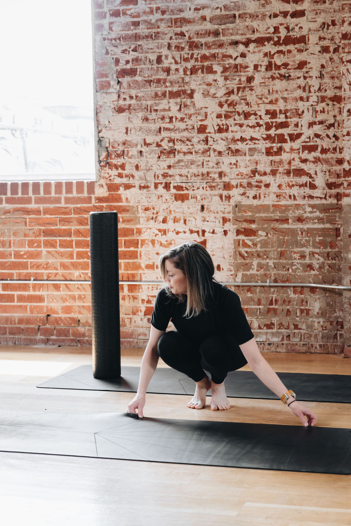 Woman in black yoga clothes adjusting her mat on a wooden floor in a studio with exposed brick wall and large window in a neighborhood wellness club in Denver.