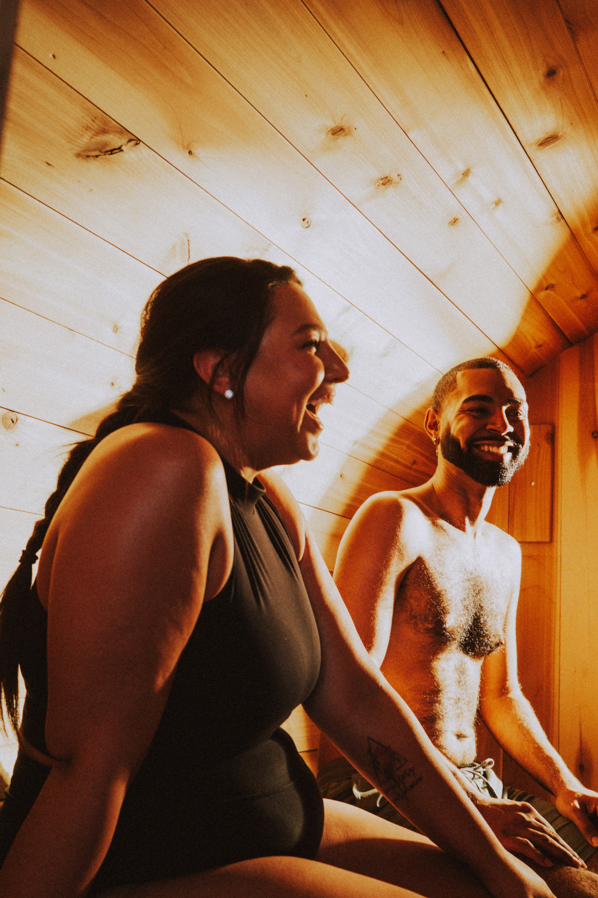 Two people smiling and laughing in a wooden-paneled traditional sauna in a neighborhood wellness club; one woman in black clothing and one shirtless man with a beard.