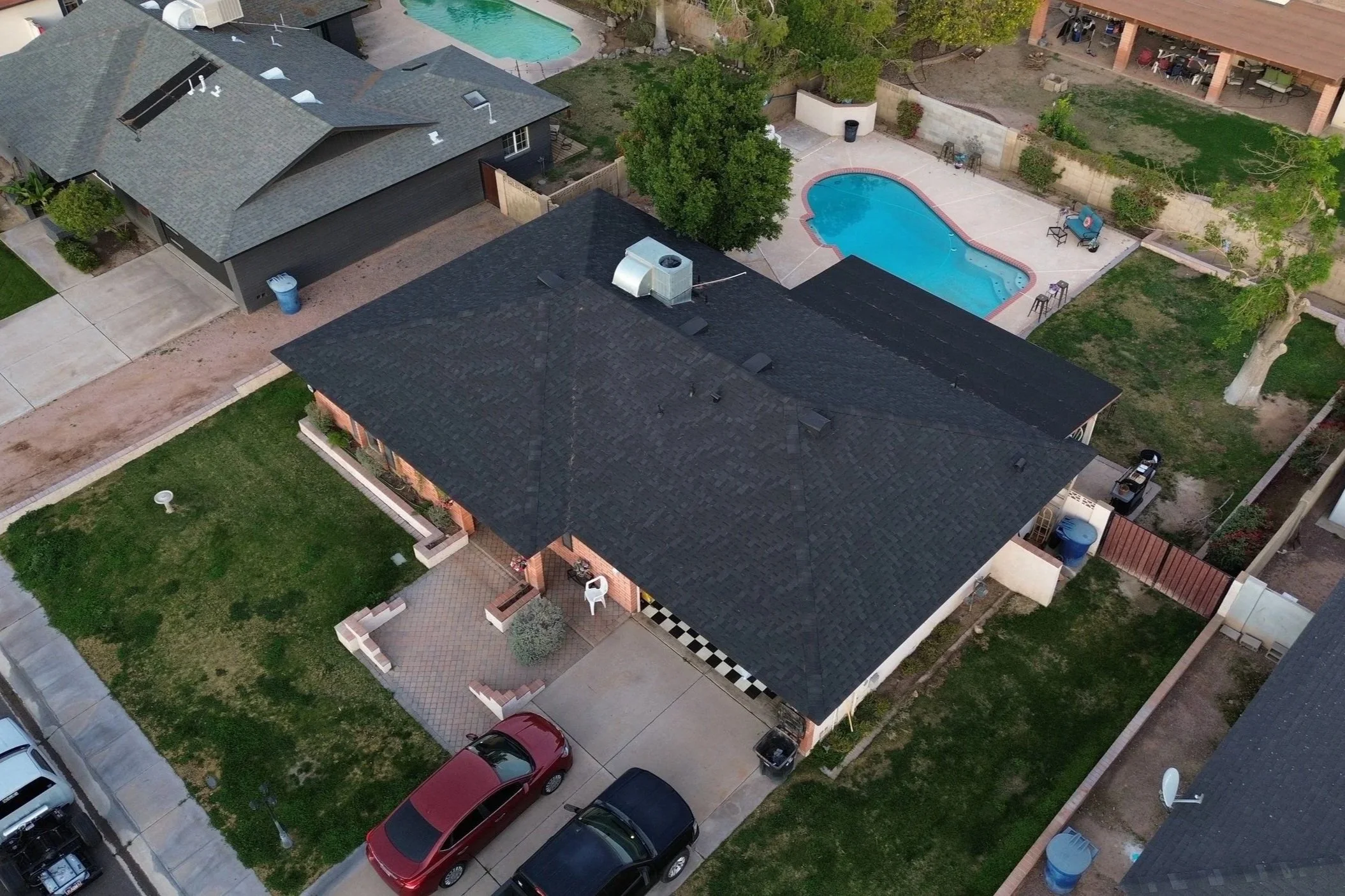 An aerial view of a suburban neighborhood showing a house with a black roof, a backyard with a swimming pool, a driveway with parked cars, and neighboring houses with yards and trees.