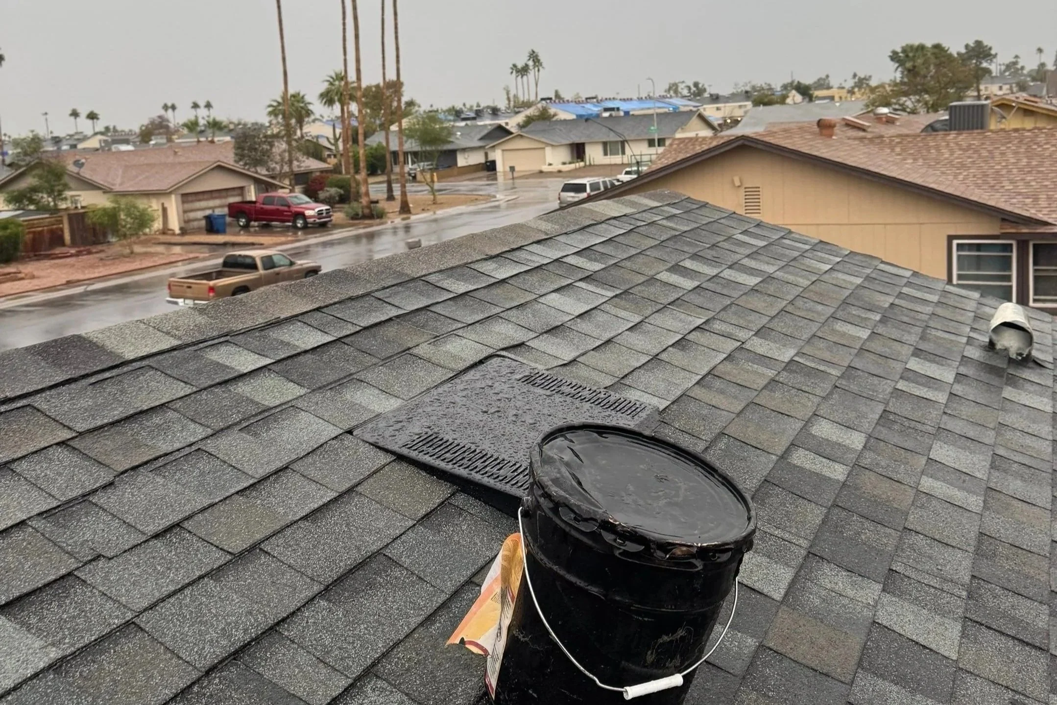Rainy day view from a roof with asphalt shingles, featuring a black bucket used for roofing work, overlooking a neighborhood street with houses and parked cars.