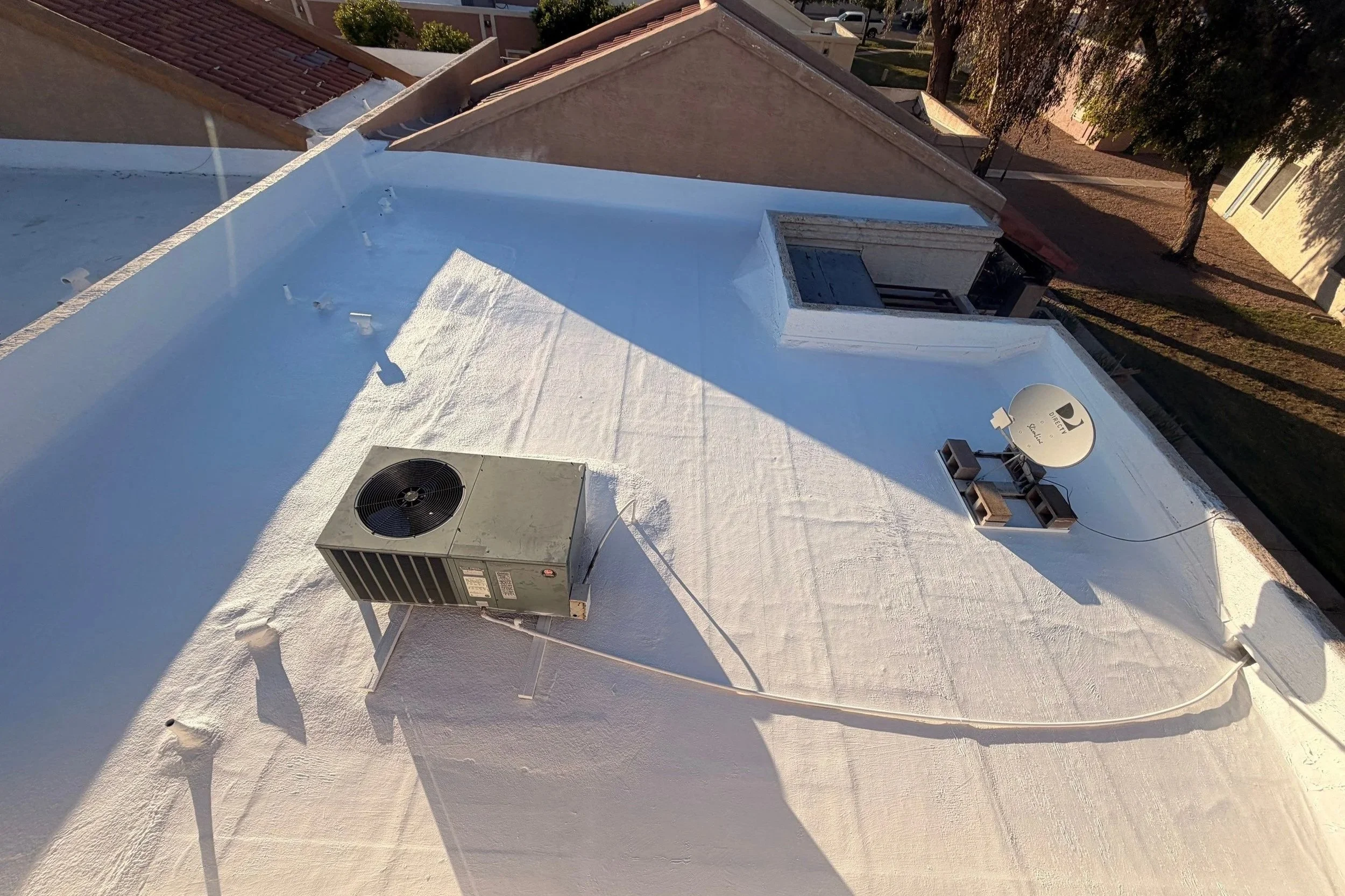 View of a flat white rooftop with an air conditioning unit, a satellite dish, and two small tables with chairs, surrounded by trees and other rooftops in the background.