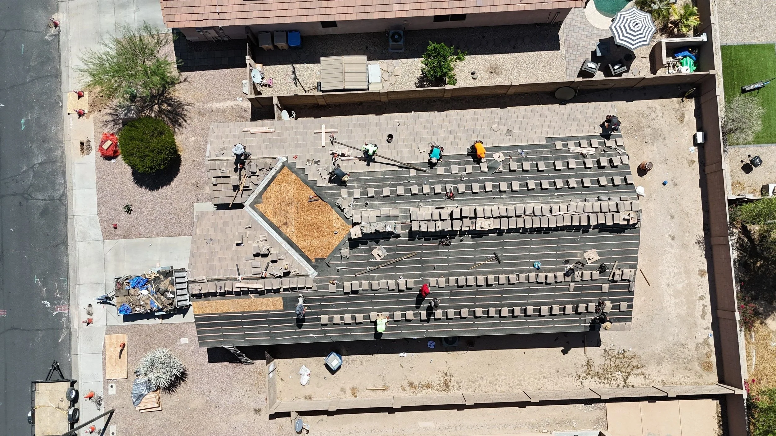 An aerial view of a construction site on a building roof, with workers installing roofing materials, scaffolding, and equipment scattered around; adjacent areas include a small courtyard with trees, outdoor seating, and gardens, enclosed by a fence.