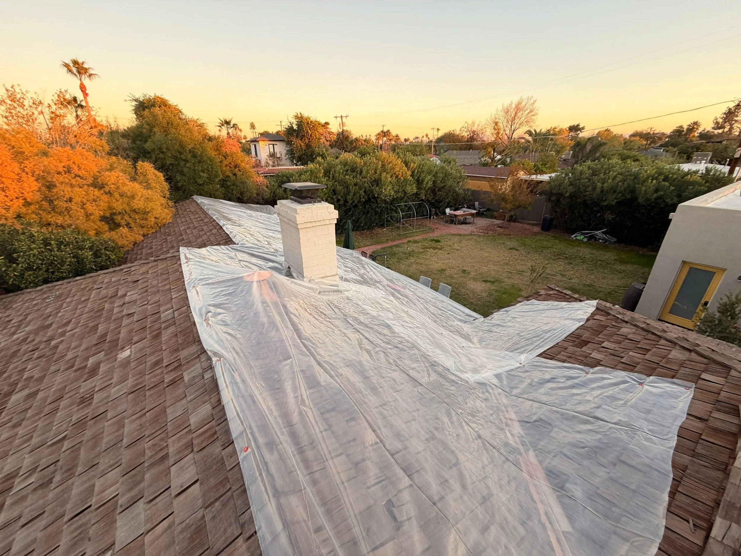 View of a house roof covered with a plastic sheet, out to a backyard with trees, a lawn, and some outdoor furniture at sunset.