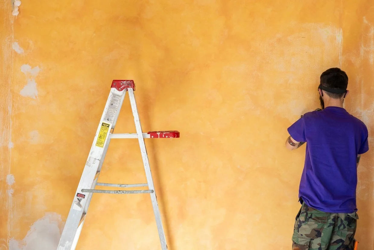 Local painter in a purple shirt working on a textured orange wall with a ladder nearby.