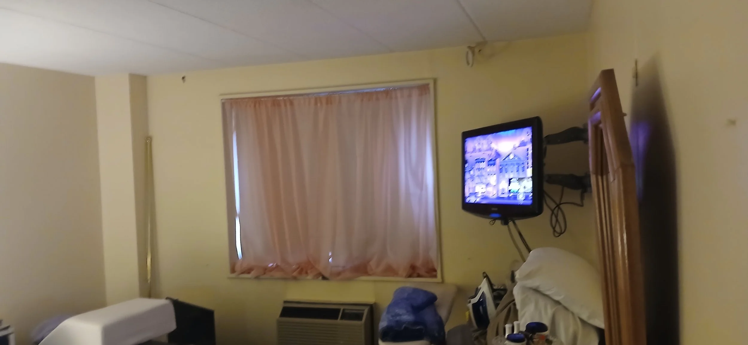 Bedroom with yellow walls and pink curtains, with a wall mounted tv that is on