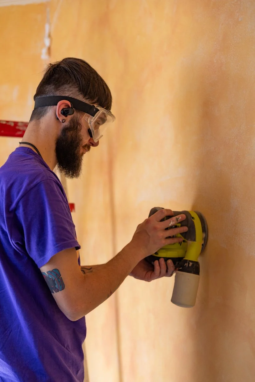Man using a power sander on a wall, wearing safety goggles and a purple shirt.