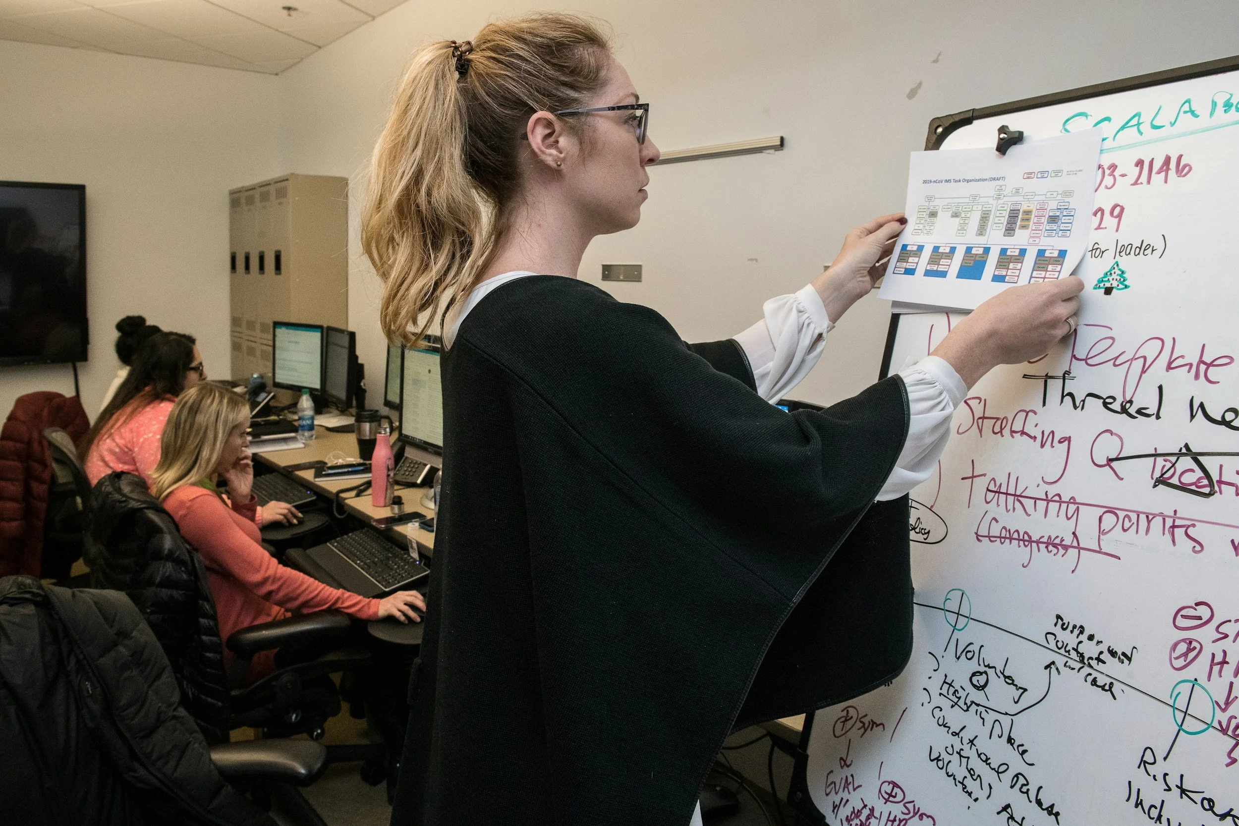 A woman with glasses and long hair in a ponytail puts up a printed organizational chart on a whiteboard. Two women are seated at desks working on computers in the background.