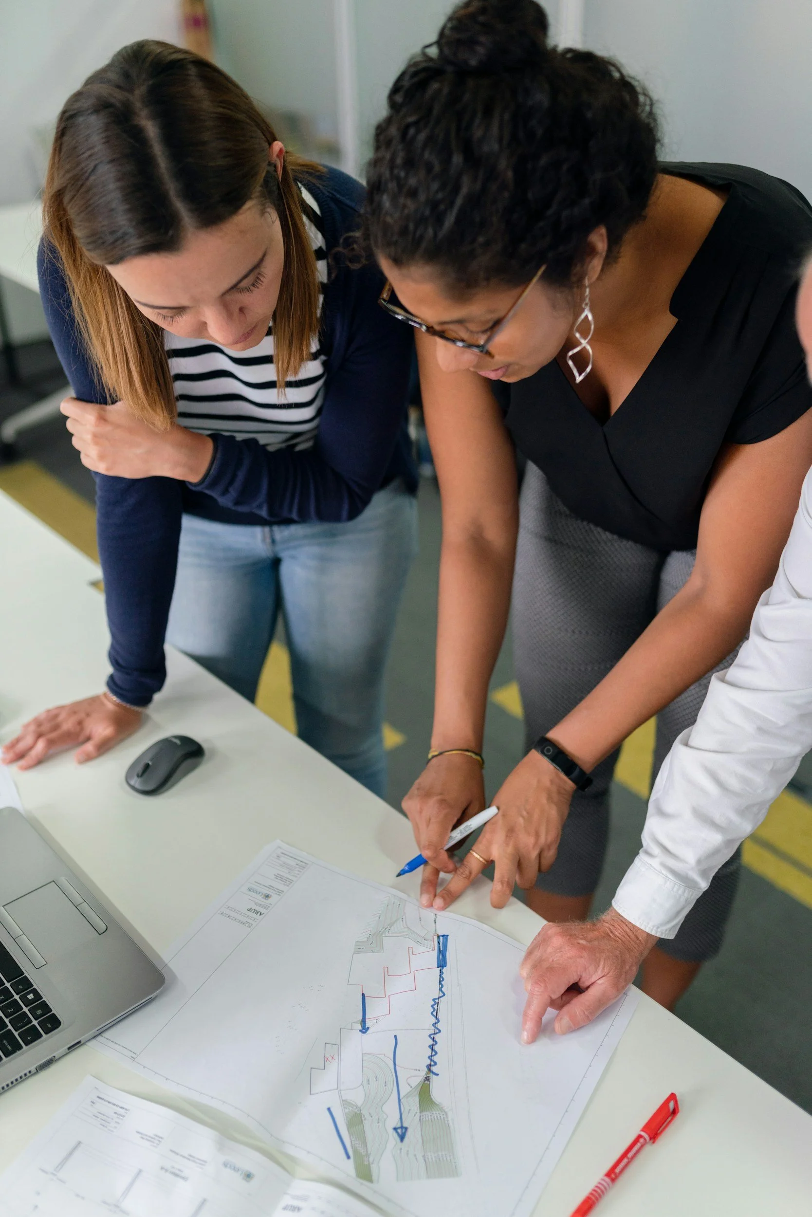 Three people are gathered around a table reviewing a landscape or architectural plan with various lines and arrows, one person is pointing at the paper with a pen.