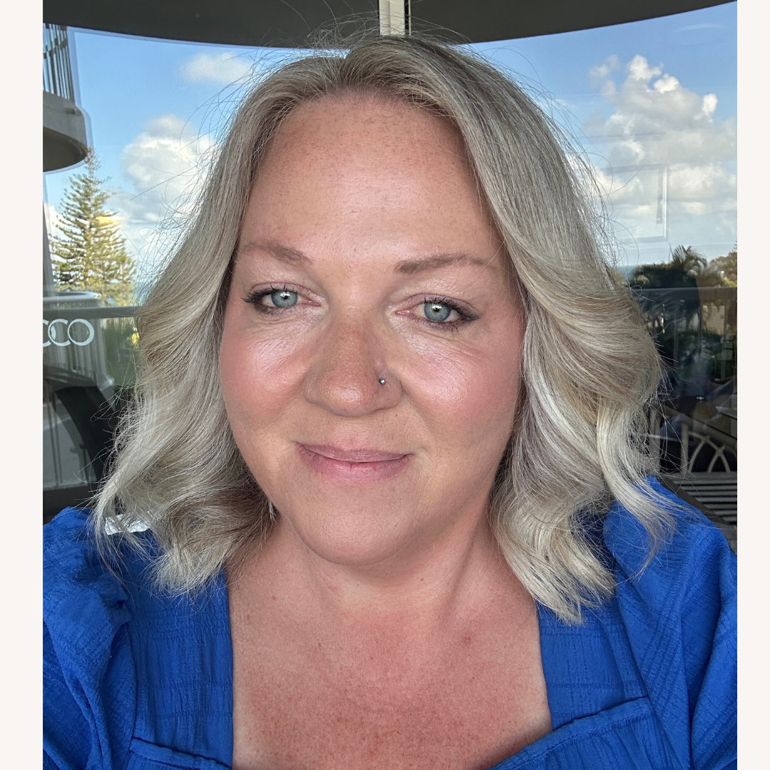 A woman with blonde, wavy hair and blue eyes smiling softly, wearing a blue top, with a sunset sky and trees in the background.