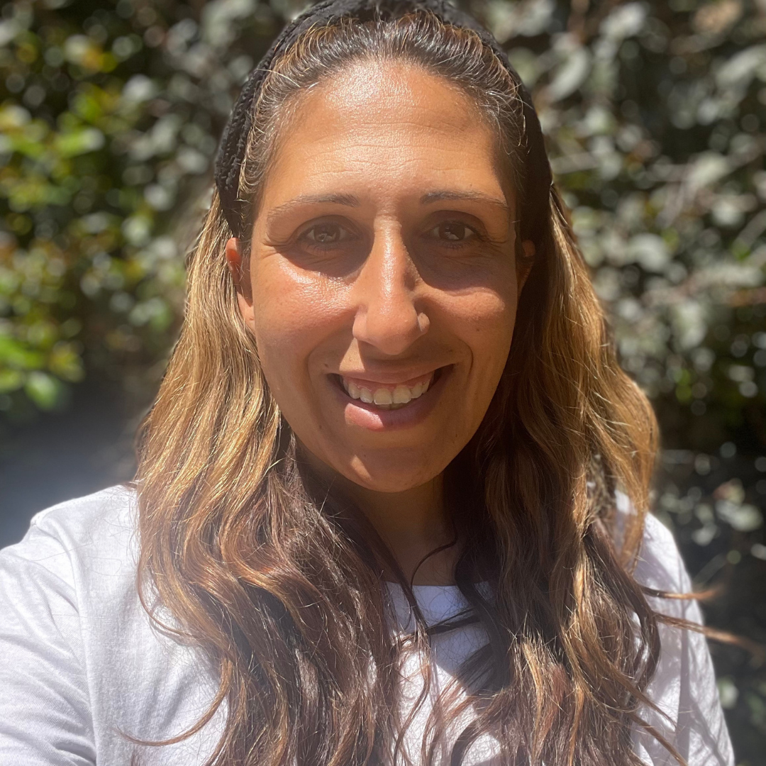 A smiling woman with wavy light brown hair wearing a white shirt, outdoors in sunny weather with greenery in the background.