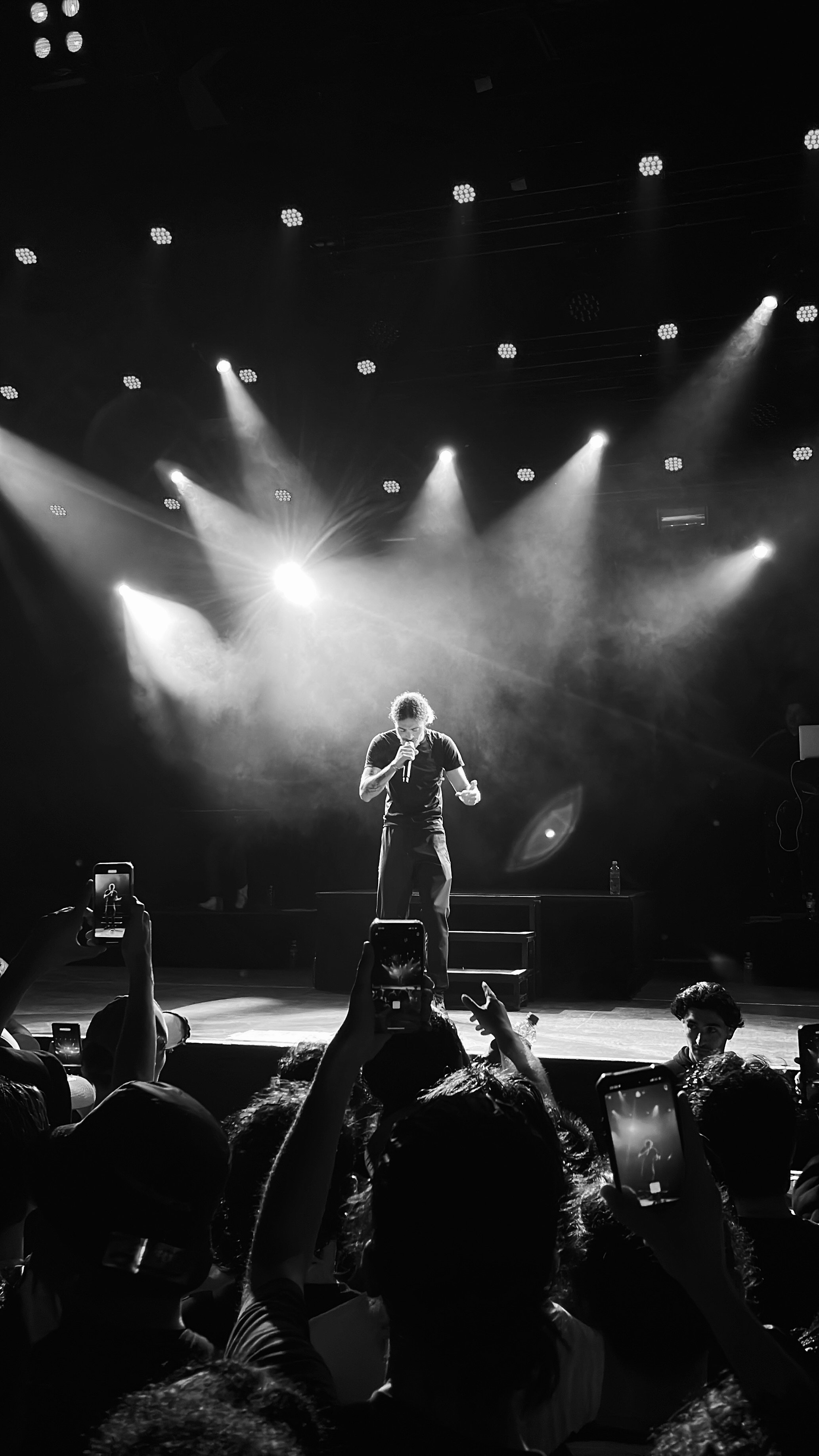 Un chanteur sur scène lors d'un concert en noir et blanc avec des lumières de projecteurs, entouré d'un public prenant des photos avec des smartphones.