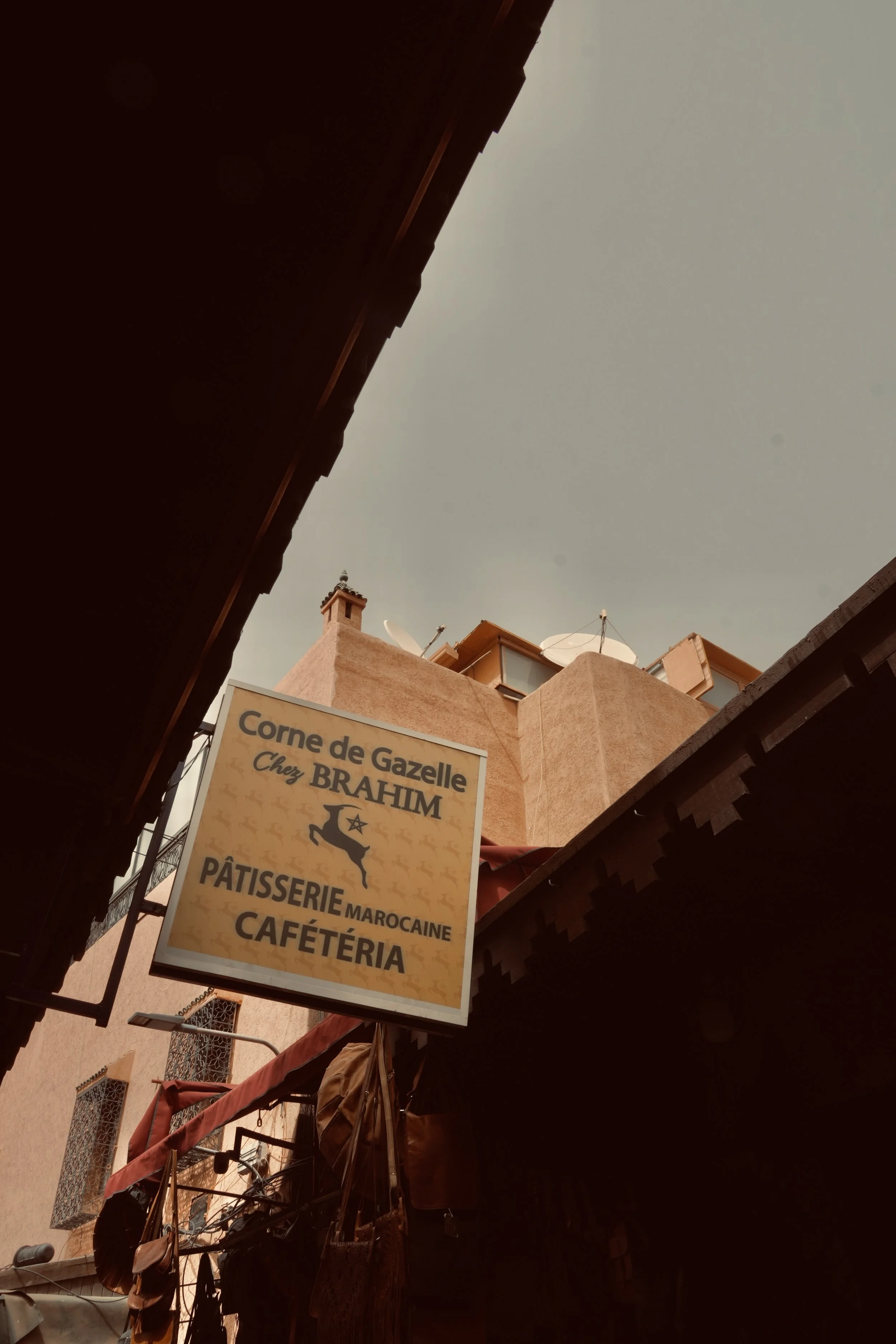 Enseigne de la boutique 'Corne de Gazelle Chez Brahim', Pâtisserie Marocaine, Cafétéria, dans une ruelle semi-ombragée avec des bâtiments aux murs couleur sable.