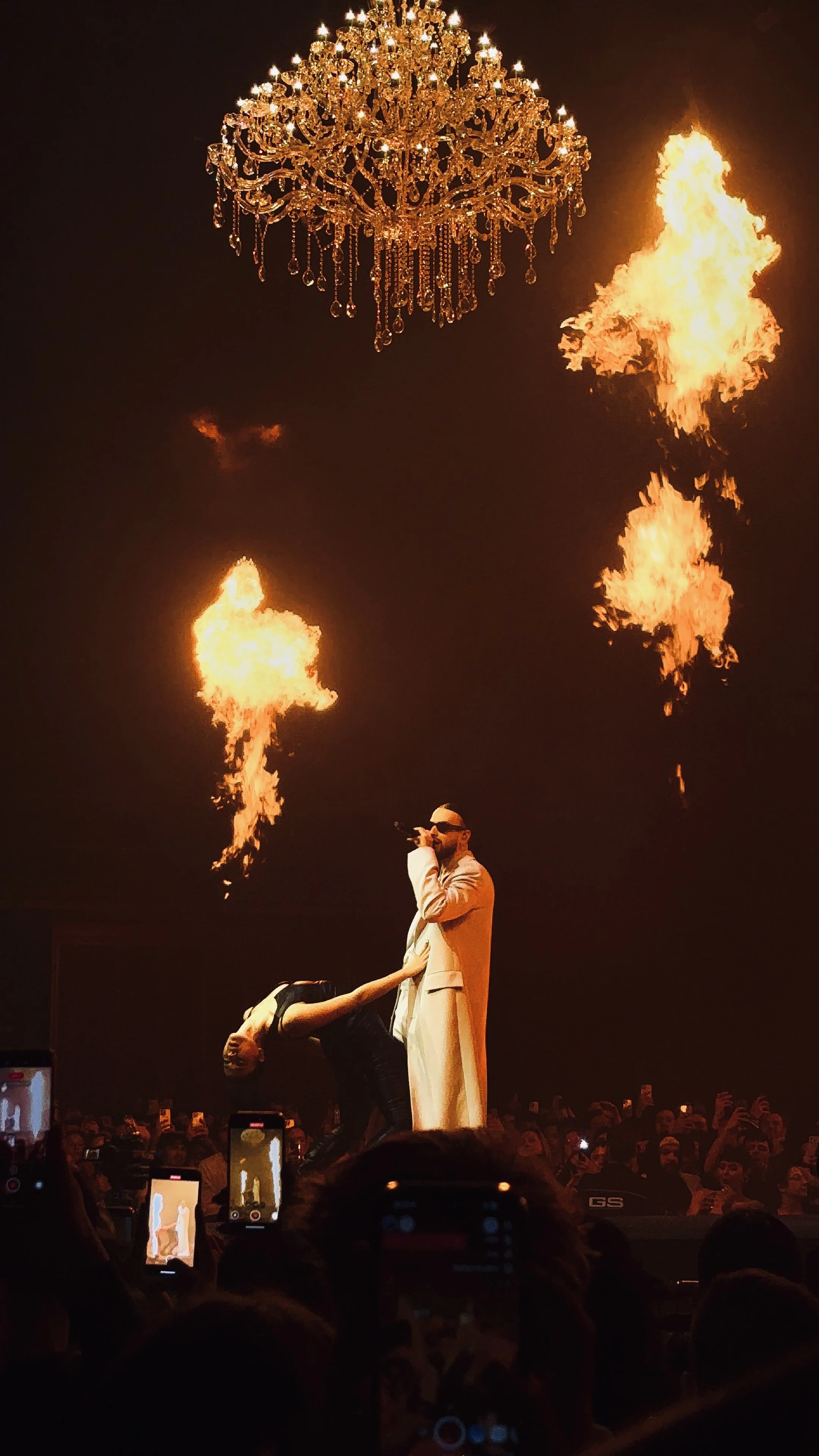 Un chanteur en scène avec un lustre au-dessus et des flammes autour, devant une foule prenant des photos avec des téléphones portables.