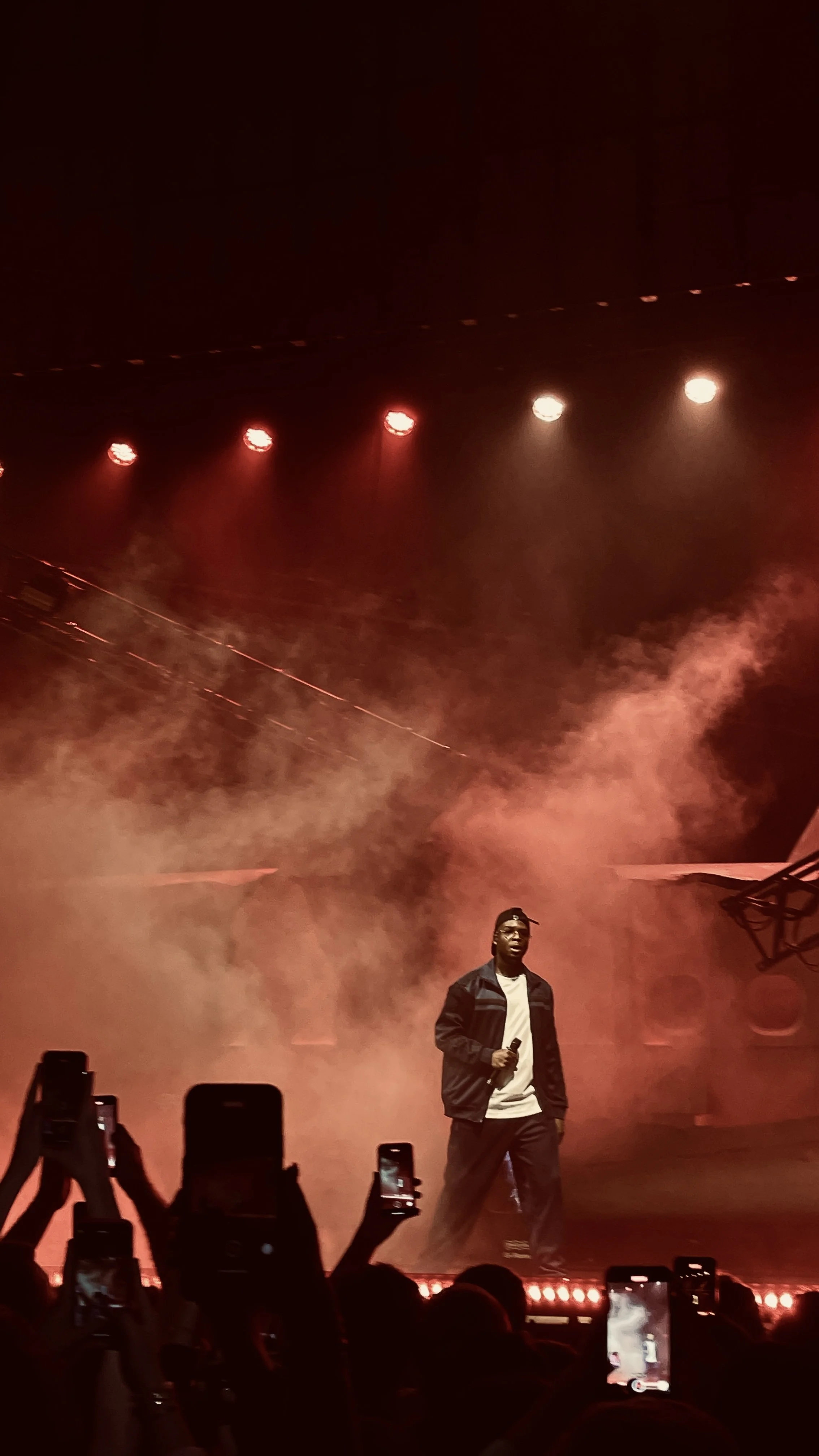 Chanteur sur scène avec micro, atmosphère fumée, lumières rouges et public photographiant avec des téléphones.