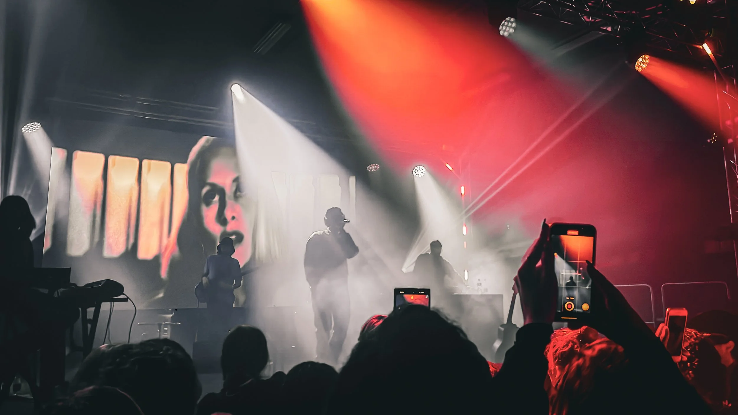 Concert avec lumières rouges et blanches, silhouettes de musiciens sur scène, écran projetant une image de visage, public avec téléphones.