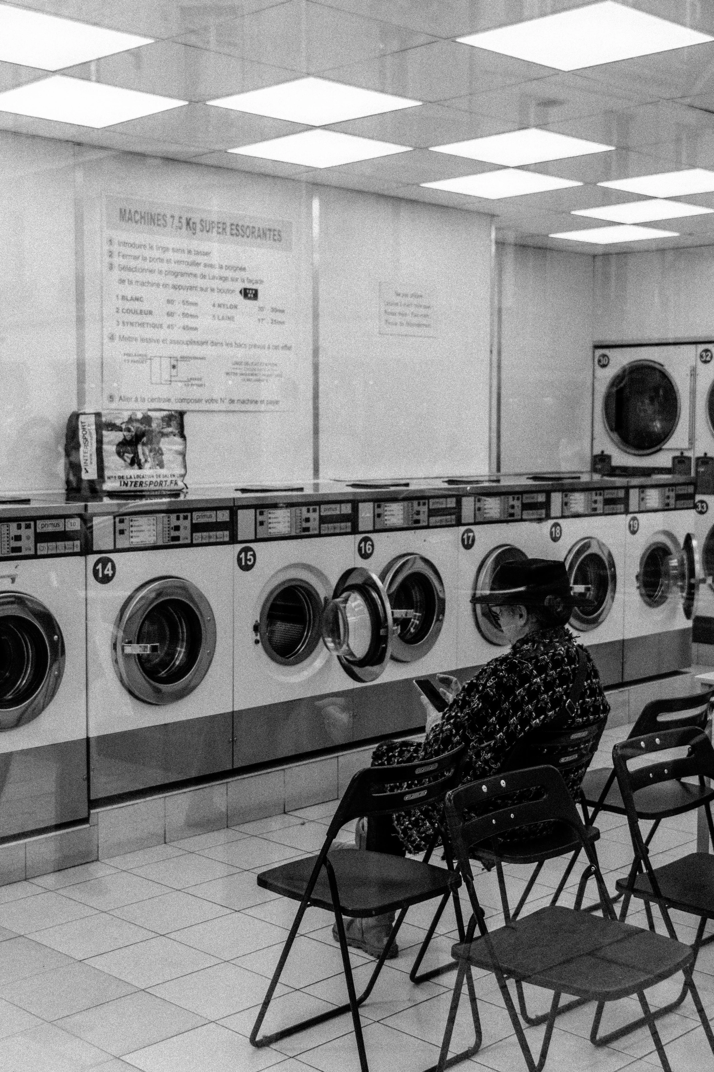 Personne assise dans une laverie automatique, entourée de machines à laver, avec des chaises alignées. Enseignes murales visibles en arrière-plan.